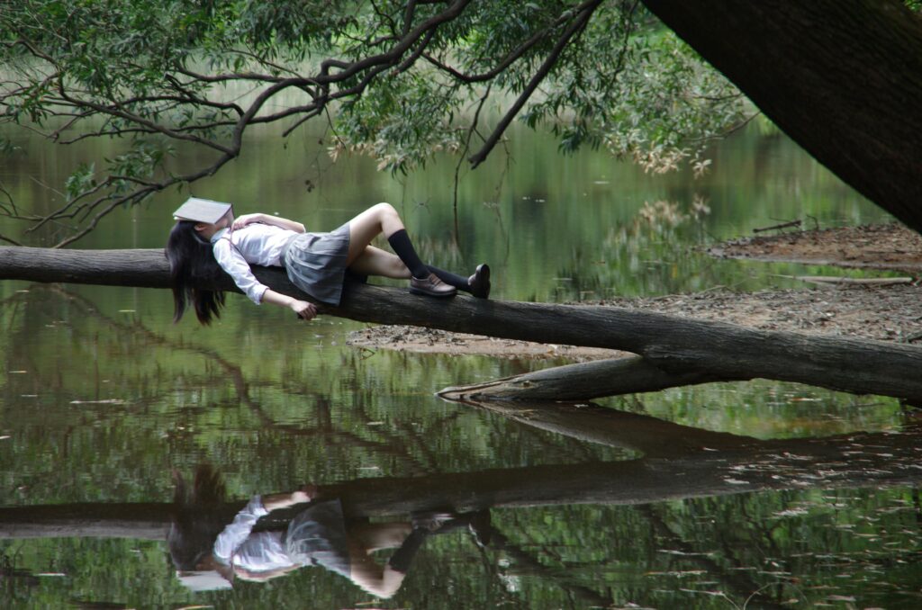 A young woman with a book on her face lies relaxing on a tree trunk by a tranquil lake.